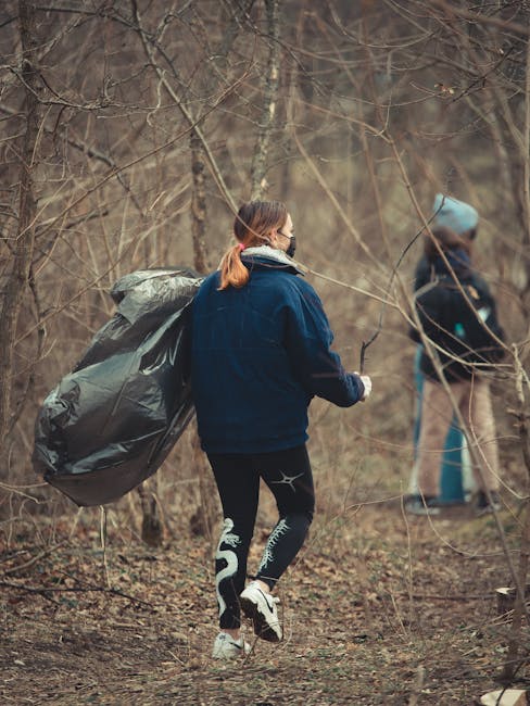 A young woman with red hair, dressed in a dark blue jacket, black leggings with white patterned designs, and white sneakers, is walking through a leafless forested area during daytime. She is carrying a large black garbage bag over her shoulder, which appears to be filled with waste or debris. In the background, another person wearing casual outdoor clothing, including a light blue hooded jacket and beige pants, is holding a trash picker and appears to be collecting litter. The environment is woodland with bare branches and a ground covered in brown leaves and twigs, suggesting late autumn or winter. The scene depicts an outdoor waste collection activity possibly related to environmental cleanup or a volunteer rubbish removal effort, highlighting the importance of responsible waste disposal and ecological preservation, akin to private rubbish removal services that focus on on-site clearance or alternative waste handling methods.