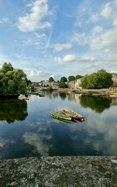 A calm river scene during daytime, with four small boats moored along the water's edge. The boats are made of wood and painted in shades of green, beige, and orange, with visible textures of weathered wood. They are positioned on a gentle slope of the riverbank, with some boats slightly overlapping. The background features a row of residential buildings with cream and white facades, partly obscured by lush green trees lining the shoreline. The sky above is partly cloudy with patches of bright blue, reflecting softly on the river's surface. The water is smooth, mirroring the sky and surrounding trees, creating a serene and quiet atmosphere suitable for outdoor leisure or private boat storage. This scene is representative of waterside properties and the type of environment where independent boat storage or private waterway access could be arranged, aligning with the context of alternative waste handling and rubbish removal services in outdoor or recreational settings.